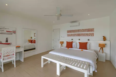 Spacious tropical bedroom with white king bed, orange pillows, wooden bench, desk, and indigenous wall art.